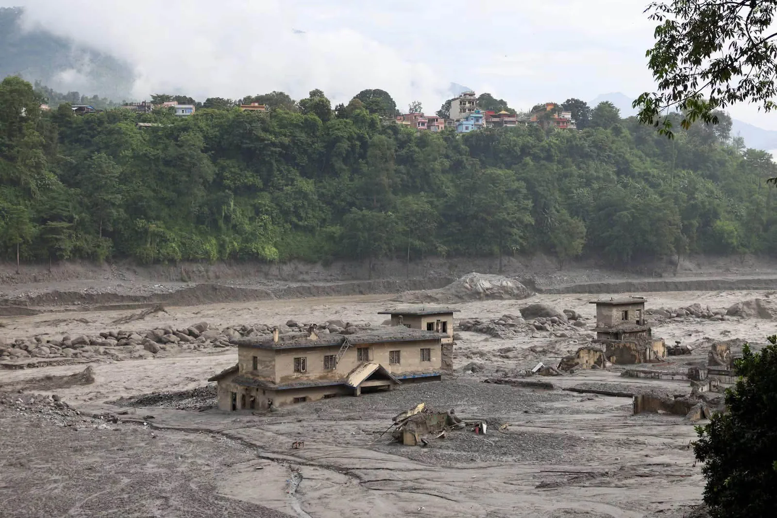 Buildings gutted and half-buried in grey silt on the Trishuli riverbank in Nuwakot after the 2026 flood, with an intact hillside settlement above