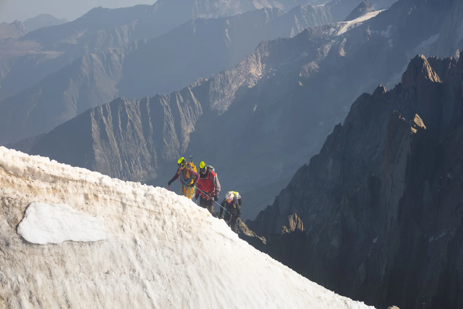 Climbers moving along an exposed snow ridge, the kind of long traverse that offers no protection for hours