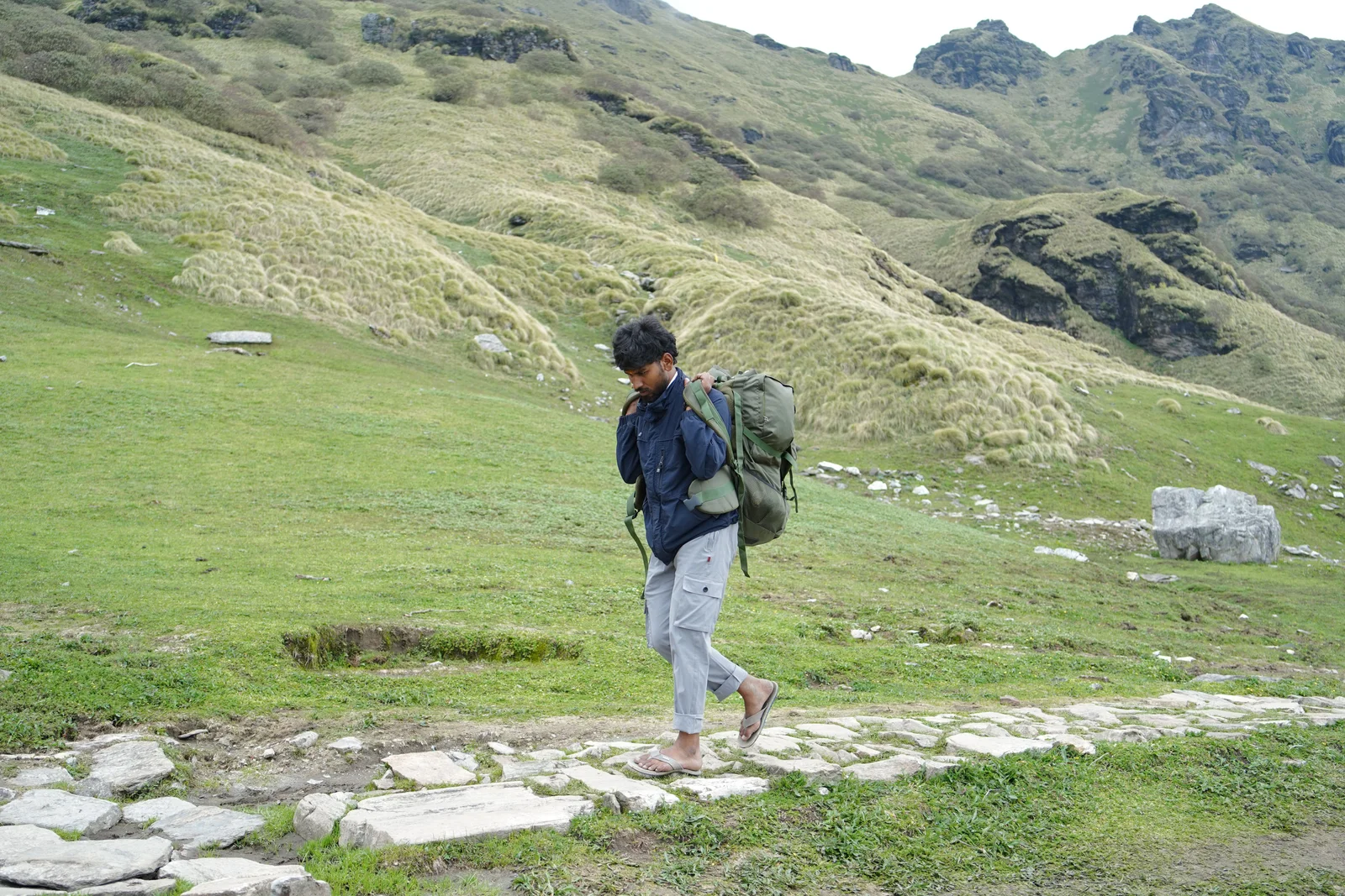 Man trekking through lush green hills in Gopeshwar, India with a large backpack, showcasing adventure and exploration.