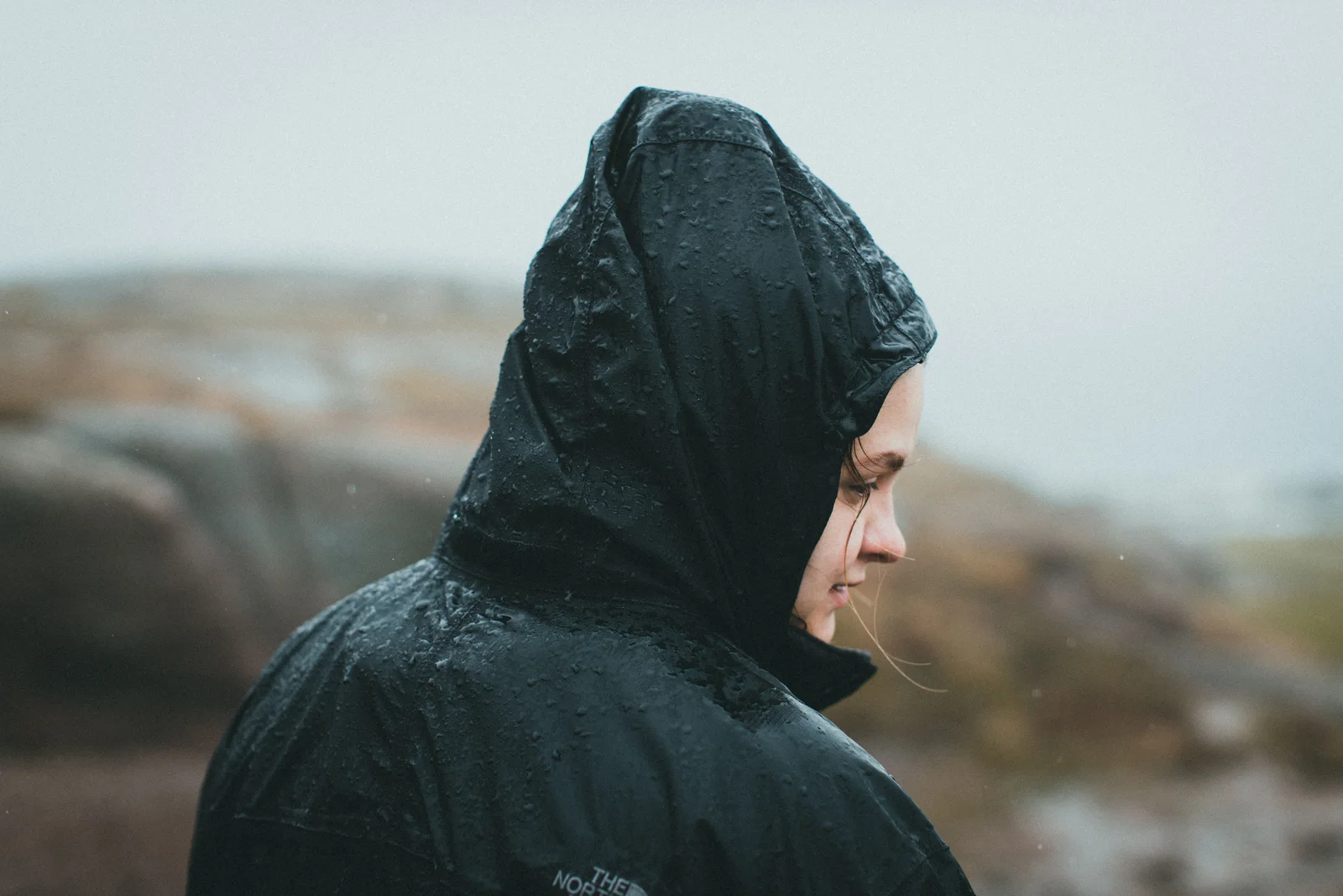 Someone in a rain jacket with the hood up in falling rain — the garment, not the fabric, is what keeps water out